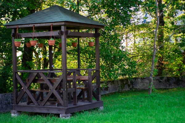 Outdoor wooden gazebo over summer landscape background. Wood brown arbour on green lawn. Summertime lush foliage pine trees blue cloudy sky sunny day nobody. — Stock Photo, Image