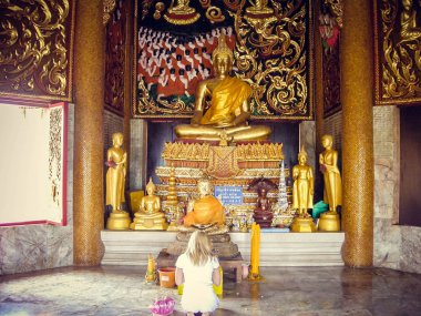 A blonde girl is kneeling in front of a statue in Thailand. Tourism. clipart