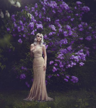 Bahar tarzı. Güzel seksi kız sarışın bahar. Çiçek açması bahar bahçe. Altın şık elbiseli Genç kız. Genç kadın Mor çiçek açan ile bahçedeki çalı