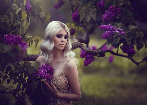 Portret van een mooie sensuele jonge blonde vrouw in het voorjaar. Bloeiende lentetuin. Jong meisje in een gouden jurk. Lente stijl. jonge vrouw in een tuin met bloeiende paarse struiken. — Stockfoto