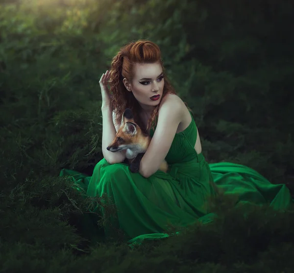 一个穿着绿色连衣裙的红头发的女孩正和一只狐狸坐在仙女的森林里。美丽的年轻女子与年轻的狐狸. — 图库照片