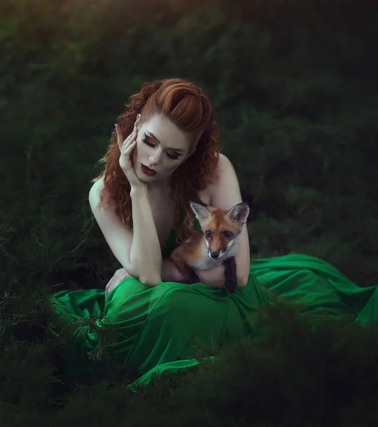 Egy vörös hajú lány egy zöld ruha ül egy Fox tündér az erdő. Gyönyörű fiatal nő, egy fiatal fox. — Stock Fotó