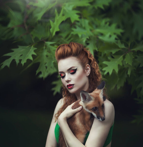 Portrait of a sensual red-haired girl in a green dress with a fox in a fairy forest. Beautiful young woman with a young fox.