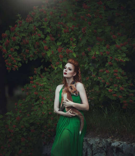 Red-haired girl in a green dress with a fox in a fairy forest. Beautiful young woman with a young fox.