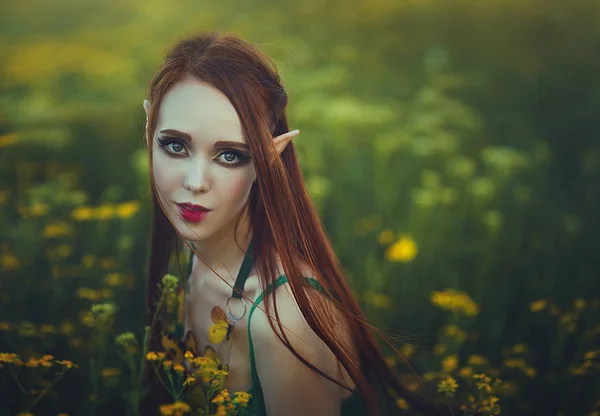 Retrato de uma menina ruiva elfo em um maiô verde posando em uma clareira de flores amarelas. Fantástica jovem com borboletas amarelas nos raios de luz . — Fotografia de Stock