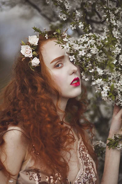 Una mujer suave con el pelo largo y rojo en un floreciente jardín de primavera. Chica sensual rojo con la piel pálida y ojos azules con aspecto inusual brillante se esconde detrás de las flores . —  Fotos de Stock