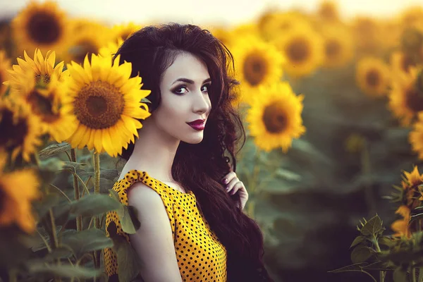 Mooie brunette vrouw in gele jurk in zonnebloemen veld bij zonsondergang. — Stockfoto