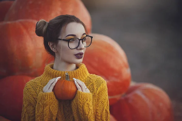 Mysig höst foto av en flicka med pumpor i en gul tröja och glasögon. Snygg ung kvinna i höst dekorationer håller en liten pumpa. — Stockfoto