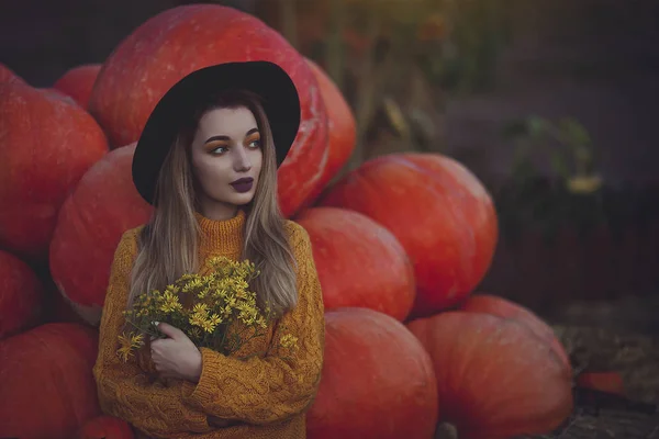 Una acogedora foto otoñal de una chica con calabazas en suéter amarillo y sombrero. Mujer joven con estilo en el paisaje de otoño, en las manos un ramo de flores amarillas . —  Fotos de Stock