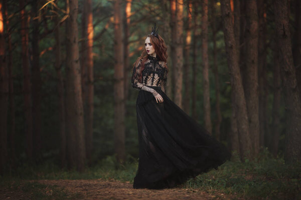 A beautiful gothic princess with pale skin and very long red hair in a black crown and a black long dress in a misty fairy forest. The costume of the dark queen. Artistic processing.