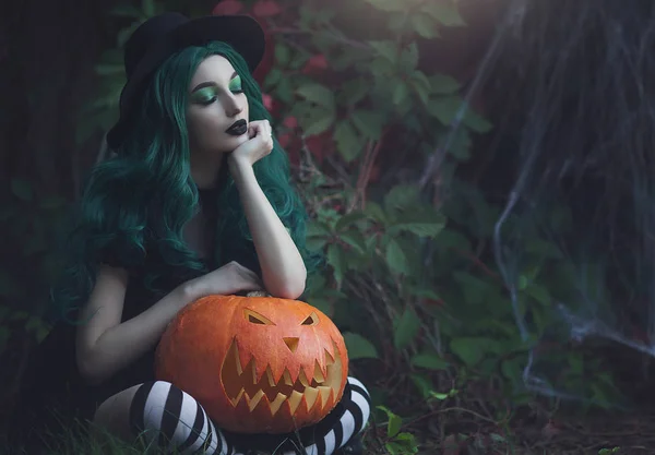 Halloween heks met groene haren houden een gebeeldhouwde eng pompoen. Mooi jong meisje in een zwarte hoed, gestreepte legging en een pak met een pompoen. Halloween-thema. — Stockfoto