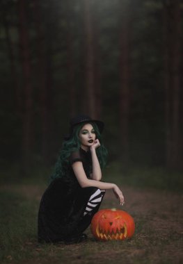 Yeşil ile Halloween cadı oyulmuş bir korkutucu Kabak yakınındaki ormanda oturuyor. Siyah şapka, çizgili tozluk ve kabak kostüm güzel ve genç birbayan. Halloween Tema.