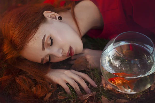 A beautiful girl with long red hair in the rays of light sleeps next to a goldfish in an aquarium. Young redheaded woman Lein on an autumn meadow. — Stock Photo, Image