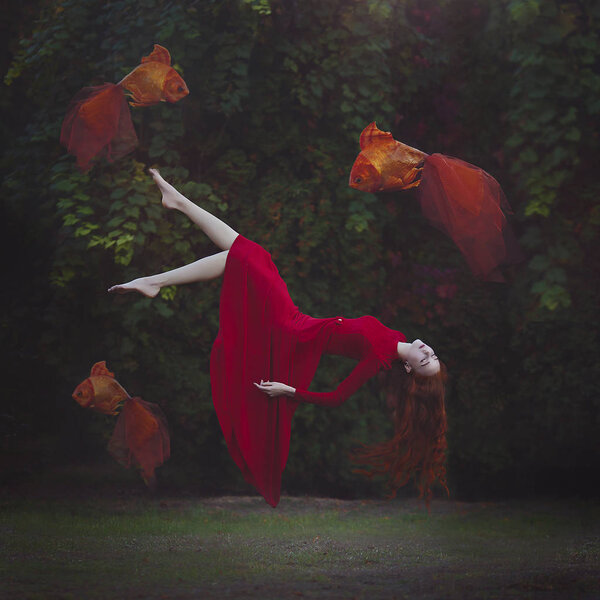 Красивая девушка с длинными рыжими волосами в красном платье левитирует над землей. Сюрреалистичное волшебное фото женщины с большой золотой рыбкой
.