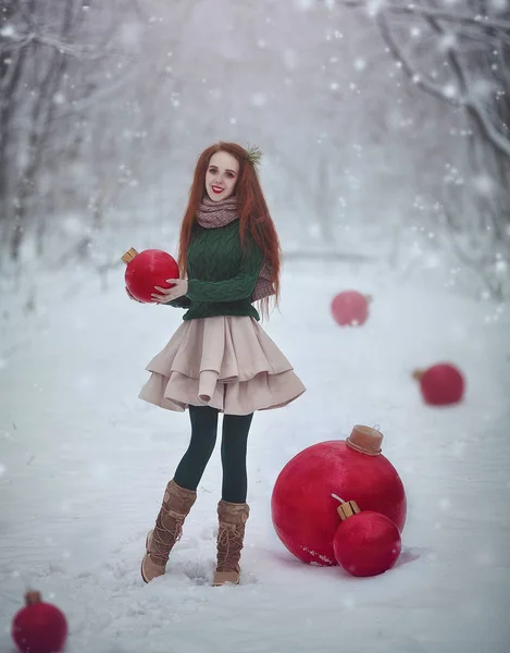 Beautiful red-haired girl like a doll with huge Christmas red balls walks in the winter fairy forest. Christmas card. Happy woman walking in snow . — Stock Photo, Image