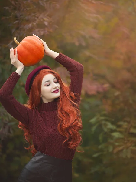 Portret van een roodharige vrouw met een pompoen in de herfstzon op de achtergrond van herfstbladeren. — Stockfoto