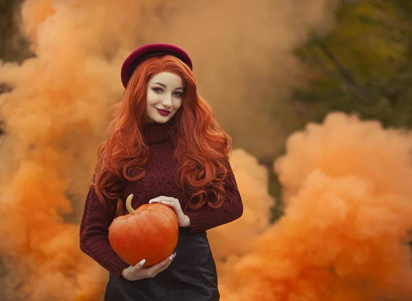 Červenovlasá holka s dýní v oranžovém kouři. Žena na oranžovém podzimním pozadí. — Stock fotografie