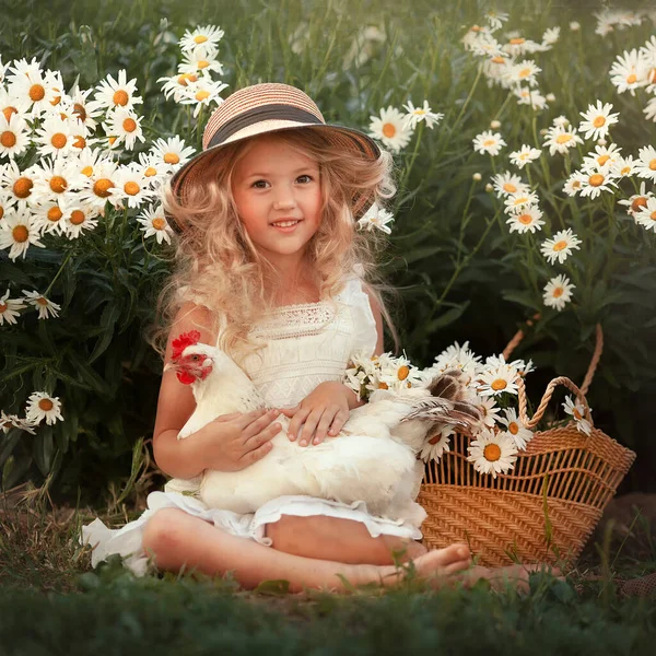 Een klein lachend meisje met een strohoed en een kip zit onder een kamillestruik. Landmeisje met een mand bloemen. Begrip "dierenbescherming". — Stockfoto