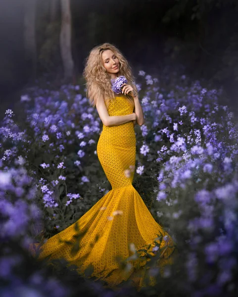 Una bella ragazza in un elegante abito giallo si trova in una foresta fiorita primavera fata in fiori viola. — Foto Stock