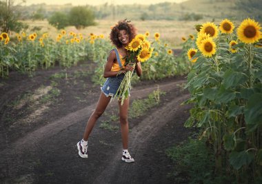 Mutlu siyah kadın ayçiçeği tarlasında yürüyor. Bir buket ayçiçeği ve kıvırcık saçlarıyla gülümseyen esmer tenli kız..