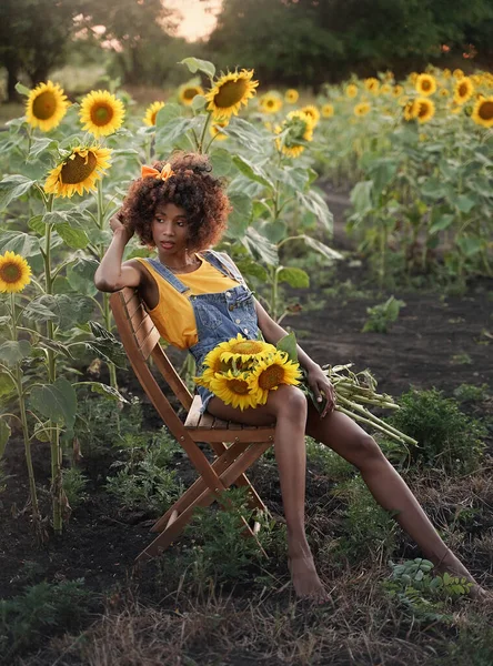 Gün batımında bir ayçiçeği tarlasında sandalyede poz veren esmer tenli genç bir kadın. Koyu tenli kız bir buket ayçiçeği ve kıvırcık saçlı.