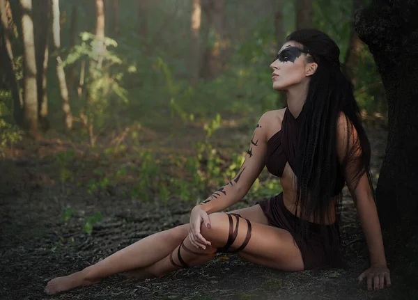 Krásná divoká amazonka s dlouhýma nohama sedí v lese. Šaman dívka s dlouhými černými vlasy a rituální make-up. — Stock fotografie