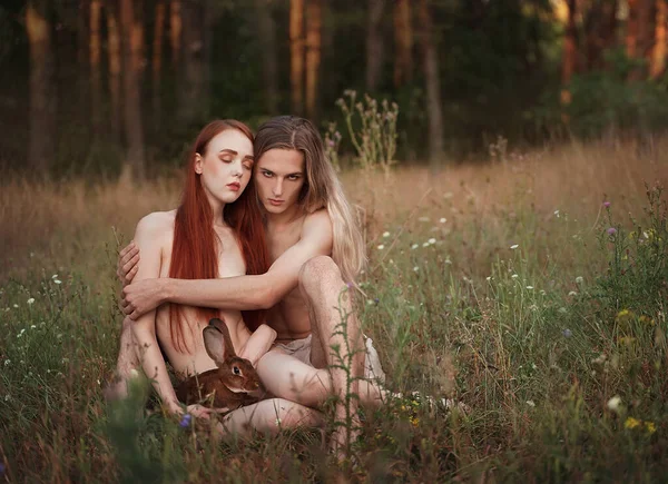 Οι πρώτοι άνθρωποι σαν τον Αδάμ και την Εύα. Όμορφος άντρας και γυναίκα με μακριά κόκκινα μαλλιά με ένα κουνέλι να κάθεται σε ένα ξέφωτο. Κοντά στο δάσος. Ενότητα με τη φύση. — Φωτογραφία Αρχείου