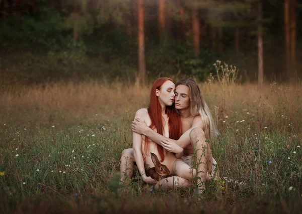 First people like Adam and Eve. Beautiful man and woman with long red hair with a rabbit sitting in a clearing. near the forest. Unity with nature. — Stock Photo, Image