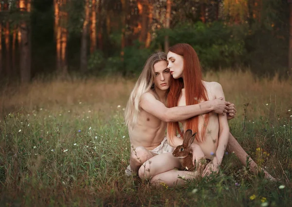 Prima gente come Adamo ed Eva. Bellissimo uomo e donna con lunghi capelli rossi con un coniglio seduto in una radura. vicino alla foresta. Unità con la natura. Foto Stock