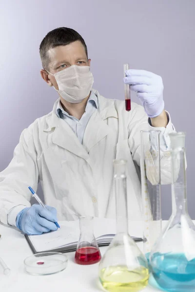 Чоловік-вчений у білій формі та масці записує свої наукові записки після перегляду аналізів крові в лабораторії. Концепція здоров'я та біотехнології — стокове фото