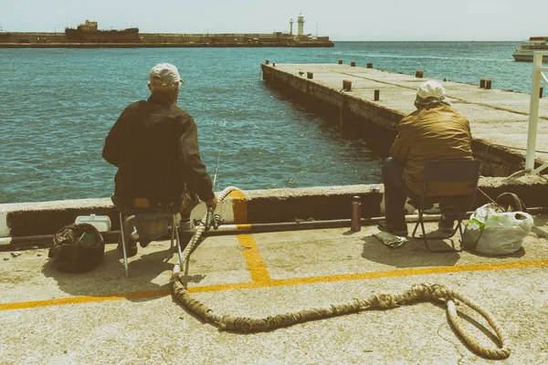 Zwei Fischer sitzen auf der Seebrücke mit dem Rücken vor dem Hintergrund einer rostigen Schiffsruine. gemildert. — Stockfoto