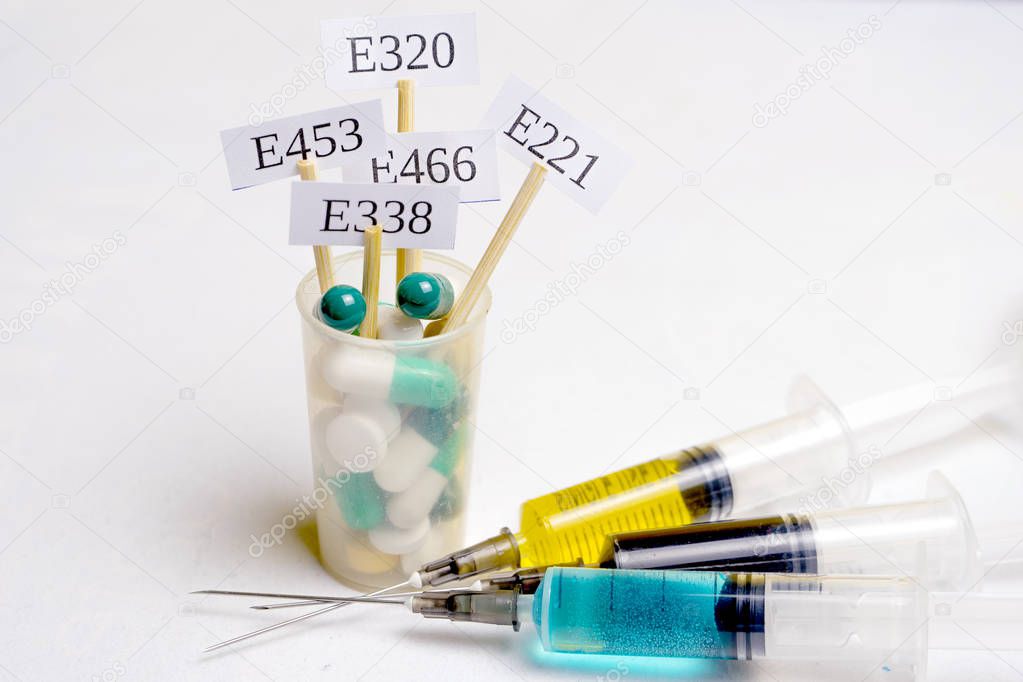 In a glass with multi-colored pills are plates with the code E-supplements. Nearby are syringes with nitrates, chemicals and additives. Harmful food additives.