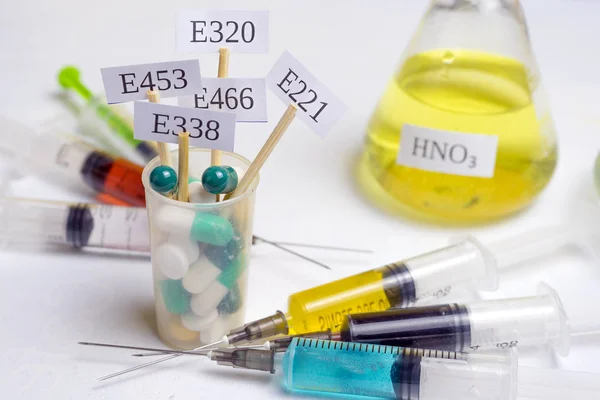 Harmful food additives. In a glass with multi-colored pills are plates with the code E-supplements. Nearby are syringes with nitrates, chemicals and additives. On the table is a flask with nitric acid