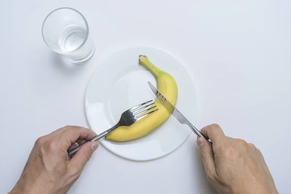 Il tema della sana alimentazione: le mani degli uomini tengono una forchetta e un coltello, una banana e piselli sotto forma di un sorriso su un piatto su un tavolo bianco — Foto Stock
