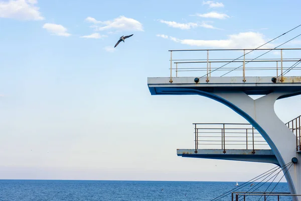 White diving board or tower against a clear blue sky. In the background the sea.