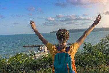 Sırt çantası olan kız denize karşı elleri ile geri duruyor.