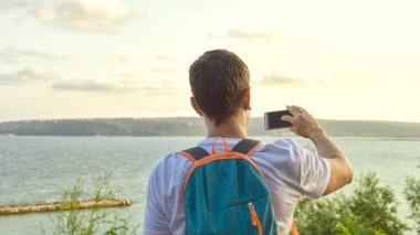 Yaşam konseptinde yolculuk. Bir erkek turist bir izleme platformu üzerinde duruyor, tatil kasabasının deniz manzarasına hayran, bir sırt çantası ile ve bir akıllı telefon üzerinde fotoğraf çekmek