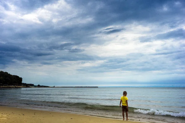 Kleiner Junge am Strand in der Nähe des Meeres, an einem bewölkten, traurigen Tag. — Stockfoto