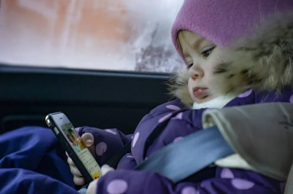 Маленькая девочка играет в телефон, сидя в машине на детском сиденье . — стоковое фото
