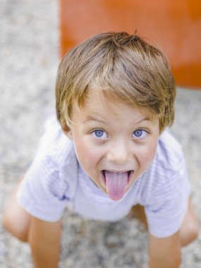 komik sarışın çocuk kameraya bakıyor ve oyun alanında kum üzerinde otururken dilini gösteren 