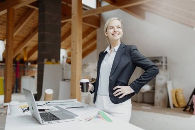 Siyah ceketli genç bayan mimar elinde kahve fincanı ve evin çatısındaki planlarla masanın yanında poz veriyor.