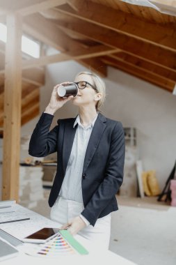 Siyah ceketli genç bayan mimar elinde kahve fincanı ve evin çatısındaki planlarla masanın yanında poz veriyor.