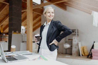 Siyah ceketli genç bayan mimar elinde kahve fincanı ve evin çatısındaki planlarla masanın yanında poz veriyor.