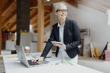 Tablet bilgisayar ve dizüstü bilgisayar kullanan genç bayan mimar elinde planlarla çatı katında masanın yanında duruyor. 