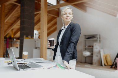 Siyah ceketli genç bayan mimar elinde kahve fincanı ve evin çatısındaki planlarla masanın yanında poz veriyor.