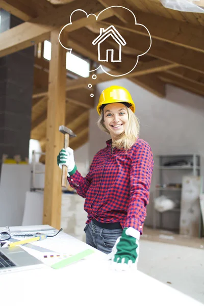 blonde woman in yellow helmet with hammer in hand standing near table with laptop and planning expansion at loft