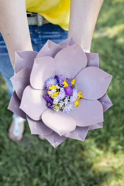 牧草地で花と美しい紫色の花のボウルを保持する女性の手 — ストック写真