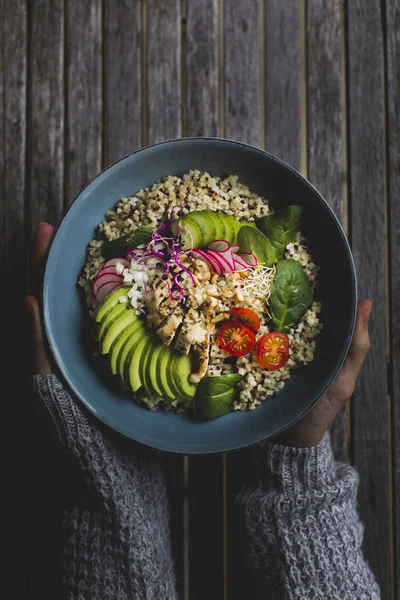 hands holding Healthy Quinoa Bulgur bowl with vegetables and chicken over wooden background