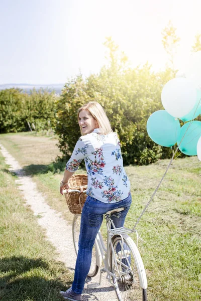 Happy woman riding bicycle with balloons on summer meadow at sunny day