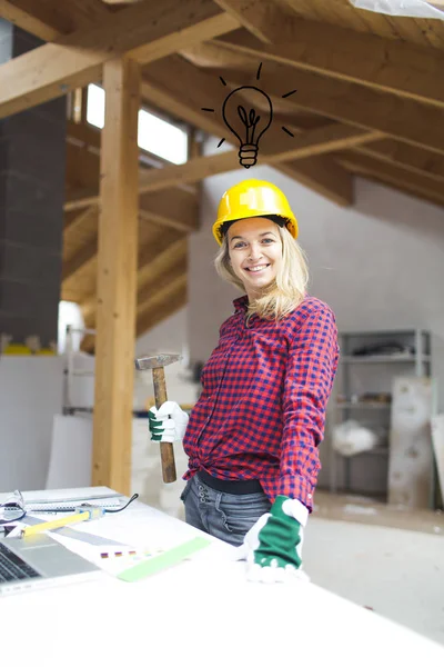 blonde woman in yellow helmet with hammer in hand standing near table with laptop and planning expansion at loft